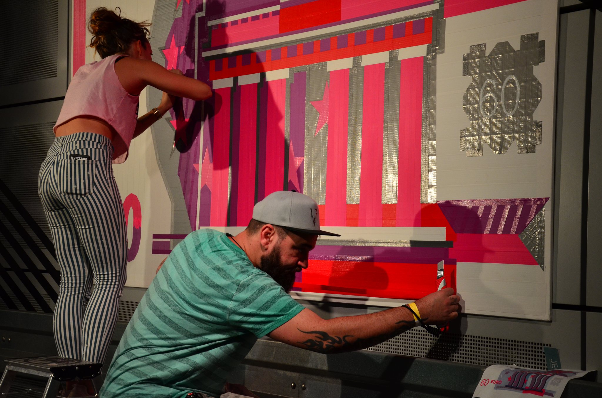 Zwei Künstler der Crew kleben ein großformatiges Tape-Art-Motiv an eine Wand